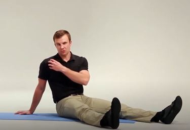 A White man is seated on a mat with his legs outstretched in a V shape. His right hand is on the ground for support, while his left hand reaches across to his right shoulder.