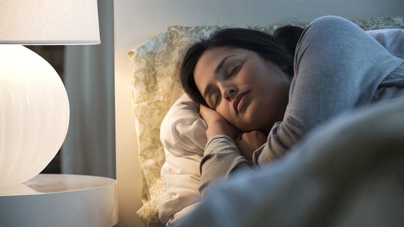 Una mujer de origen hispano/latino iluminada por una lámpara duerme en una cama.