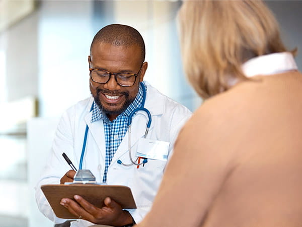 Un médico de raza negra que sonríe toma notas mientras habla con su paciente femenina de edad avanzada.