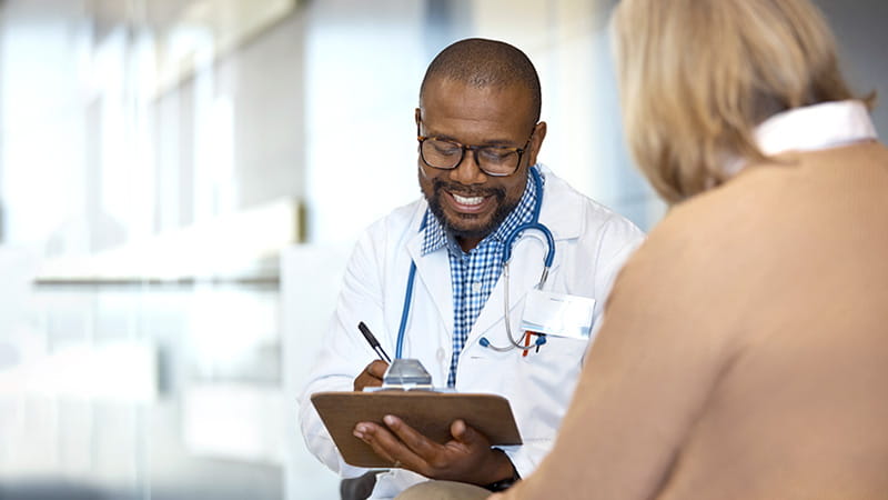 A smiling Black doctor is taking notes while talking with his senior female patient.